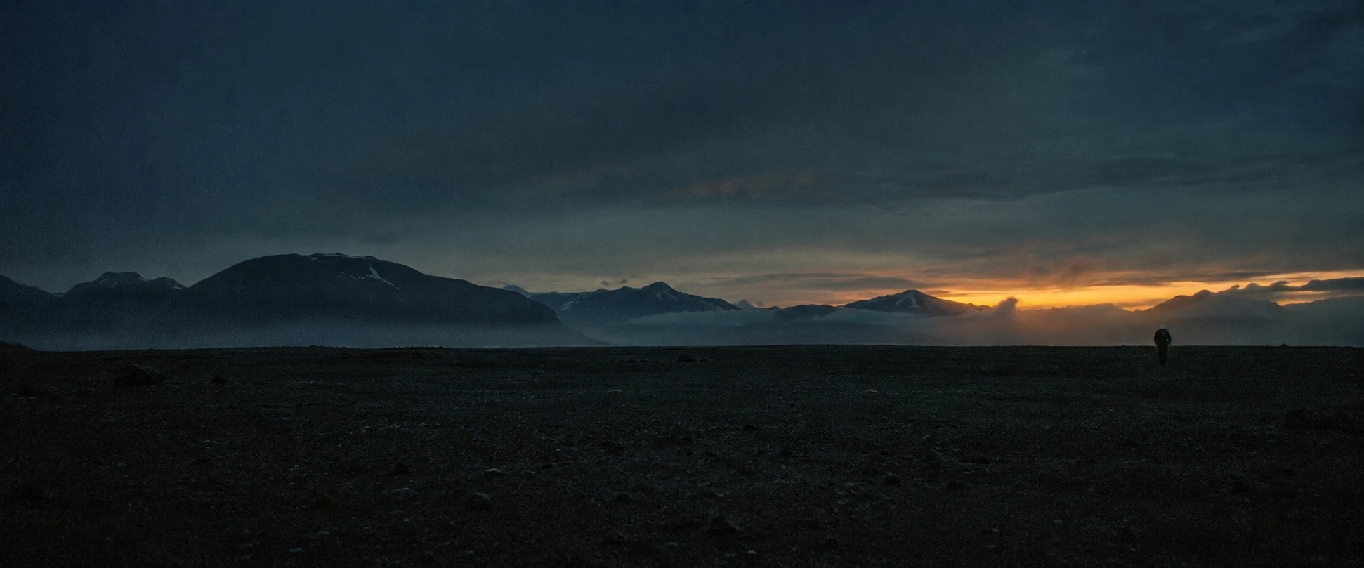 A lone figure crosses a black sand landscape beneath a sliver of amber light.